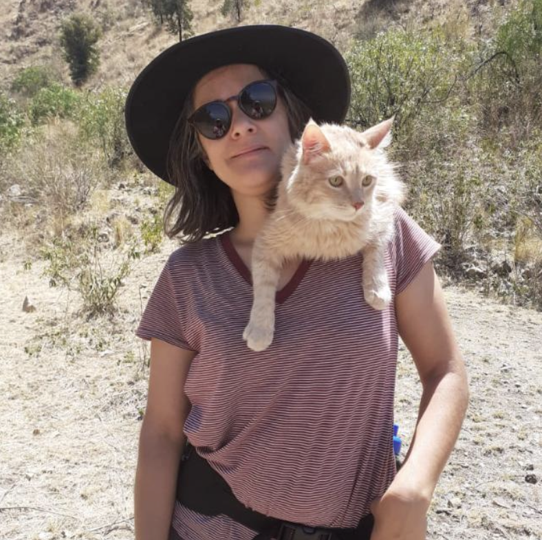 Woman in a striped shirt, wearing a wide-brimmed hat and sunglasses, holding a light-colored fluffy cat with amber eyes in an outdoor setting with dry grass and bushes.
