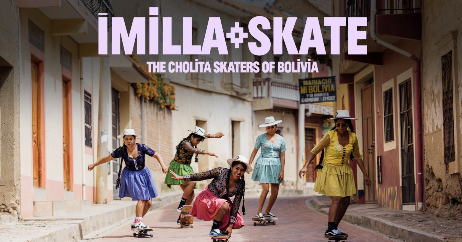 Six women skateboarding down a colorful street in Bolivia, wearing vintage dresses and wide-brimmed hats.