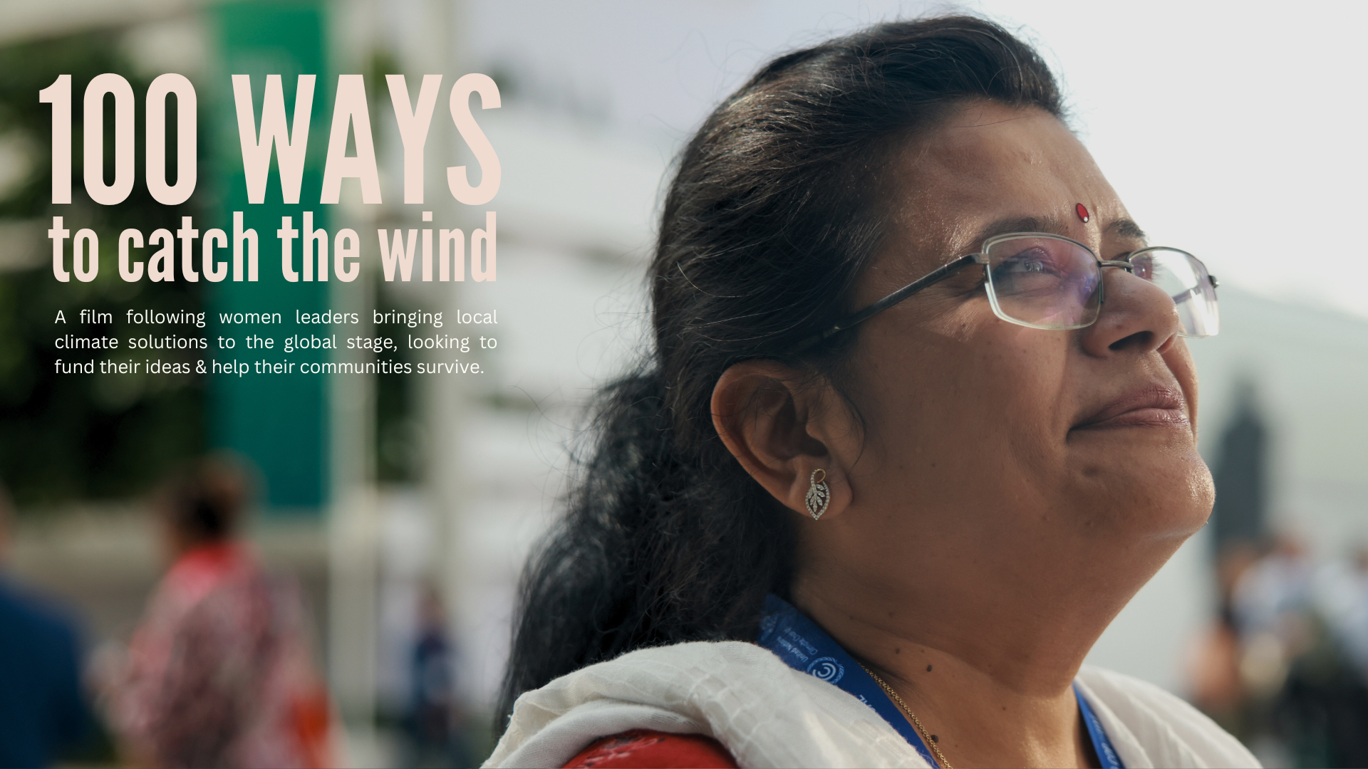 Close-up of a woman wearing glasses and earrings, looking outdoors with a slight smile, blurred background with people at an event.