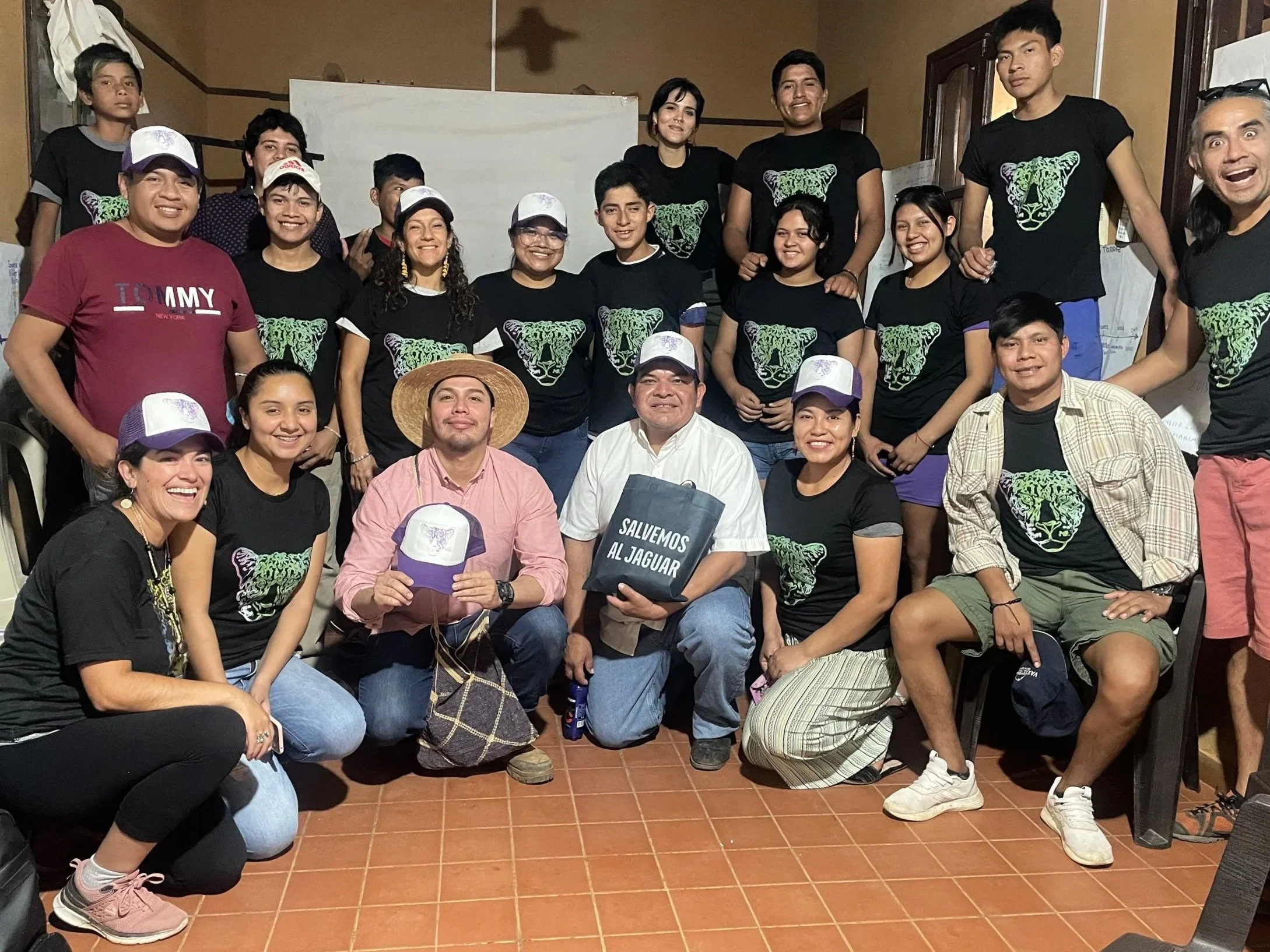 A diverse group of people smiling and posing together indoors, with some holding bags and wearing black shirts with a green tiger graphic.