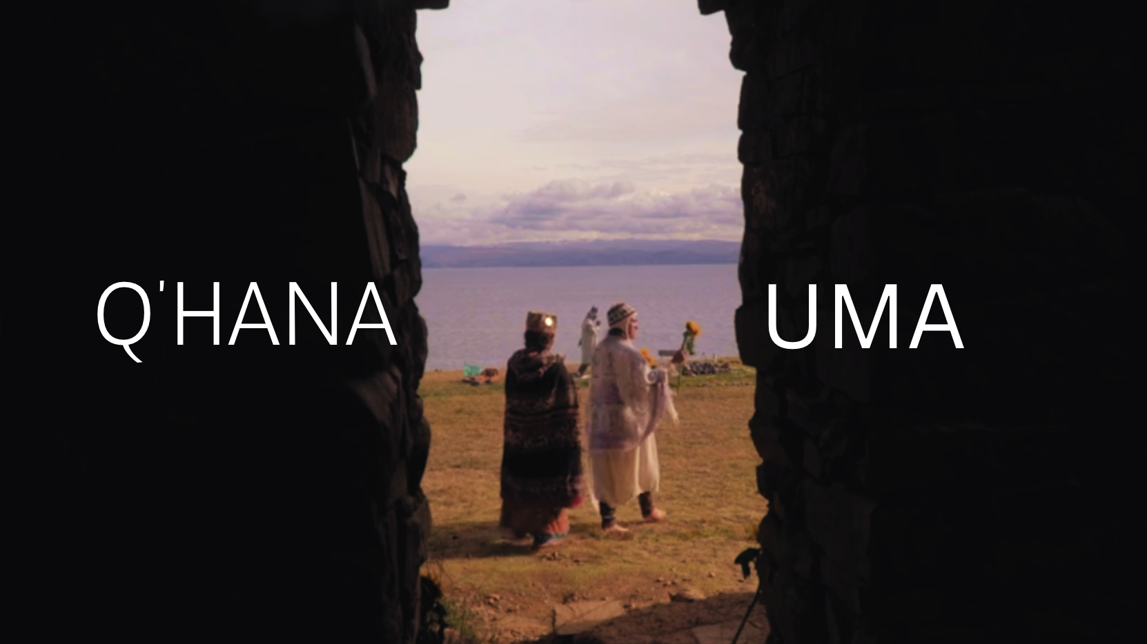 Two women dressed in traditional clothing and headgear walk near a body of water, holding flowers, as viewed through an opening in a stone structure. The background shows a cloudy sky and distant land across the water.