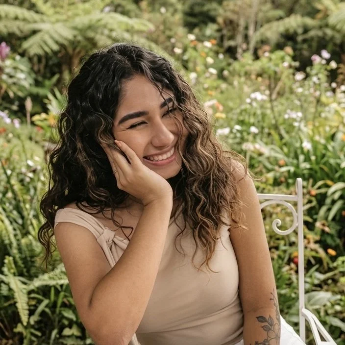 A young woman with curly brown hair, smiling and holding her face with one hand, in a lush garden.