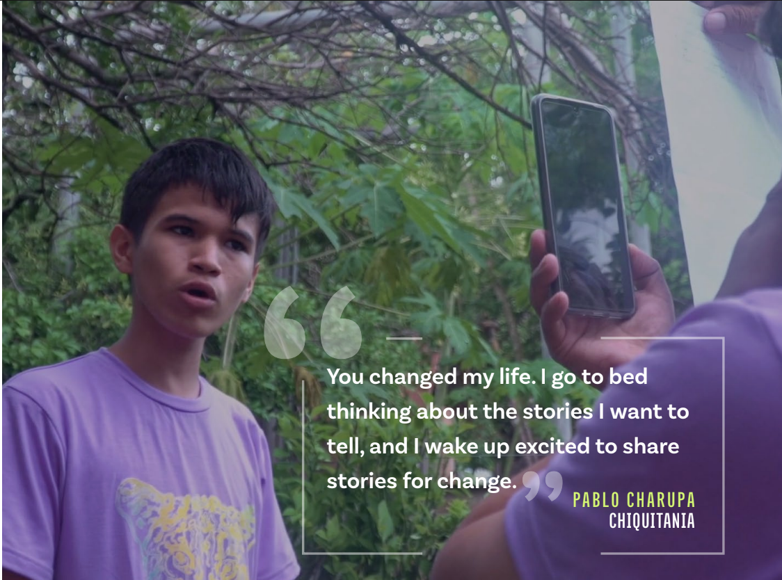 People sitting on green plastic chairs watching a movie projected onto an outdoor screen, under a thatched roof at night.