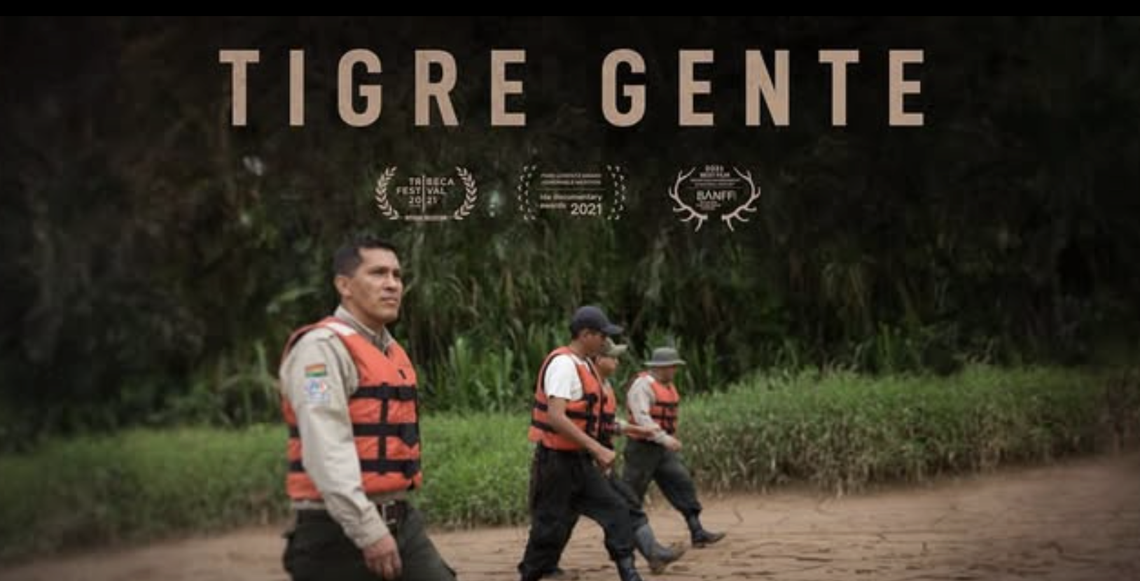 Group of four men wearing life jackets walking along a dirt path in a lush, green jungle or forest.