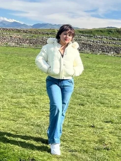 A young woman with brown hair and earrings looks upward with a playful expression against a plain wall background.