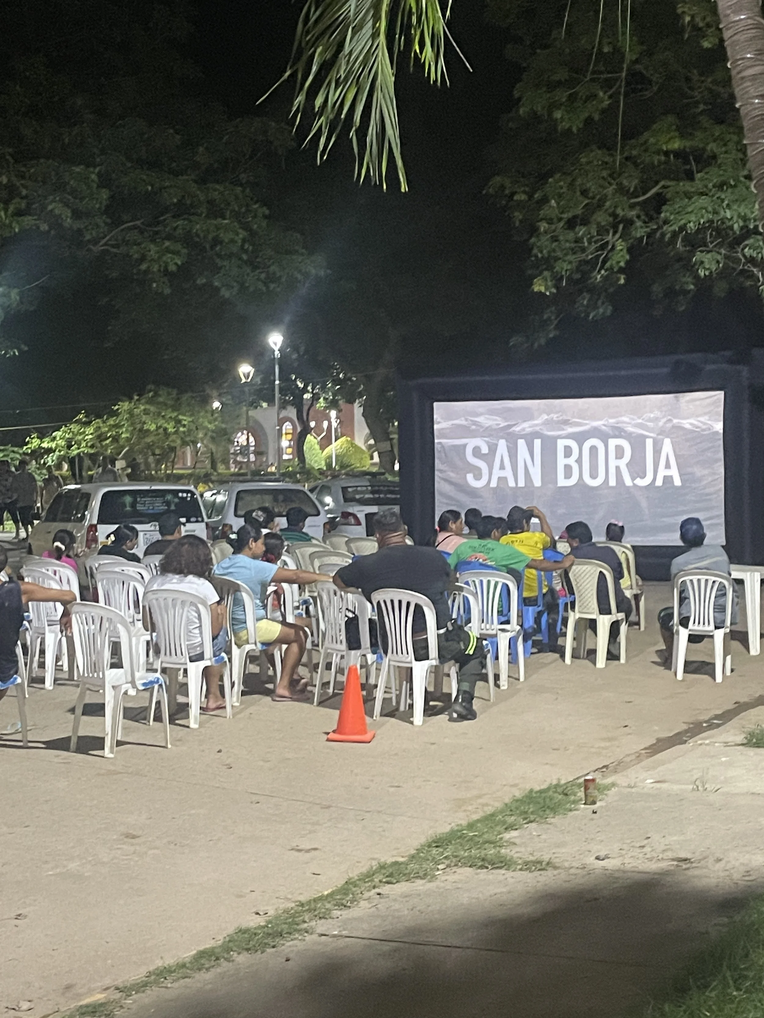 People attending an outdoor event at night, seated on bright green chairs, watching a large screen displaying mountains and a sky with clouds.
