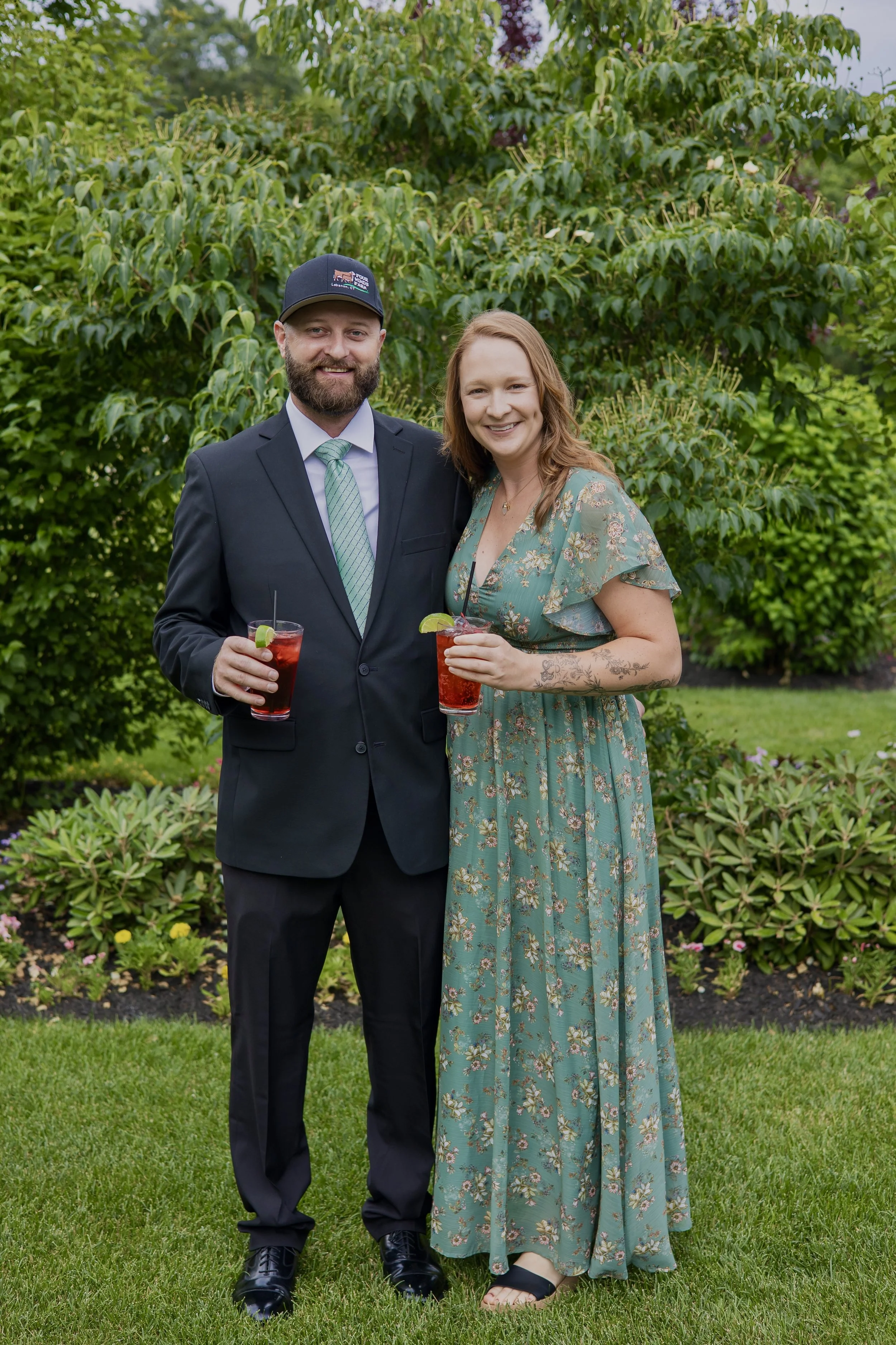 A man and woman standing outside on grass, holding drinks with lime slices, smiling at the camera. The man is wearing a navy suit with a light green tie and a black cap, which has an embroidered logo. The woman is dressed in a long, floral dress with short sleeves and sandals, with tattoos on her left arm. Behind them are green bushes and trees.