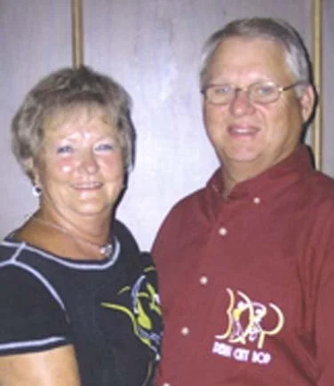 A smiling older woman in a black shirt with white and yellow accents standing next to a smiling man in a red polo shirt with glasses. They are inside a room with plain walls.