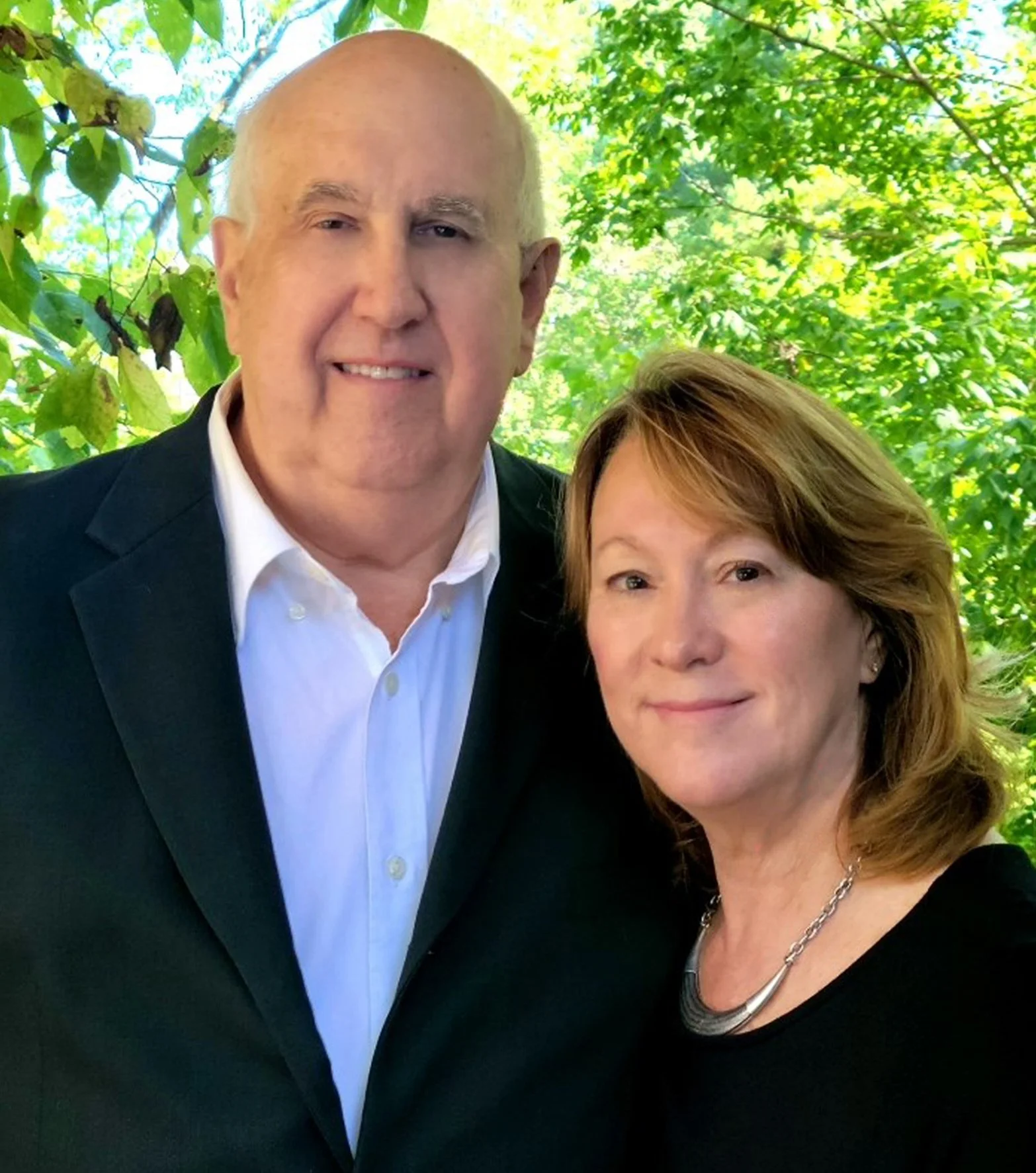 A man and a woman taking a selfie outdoors with green trees in the background.