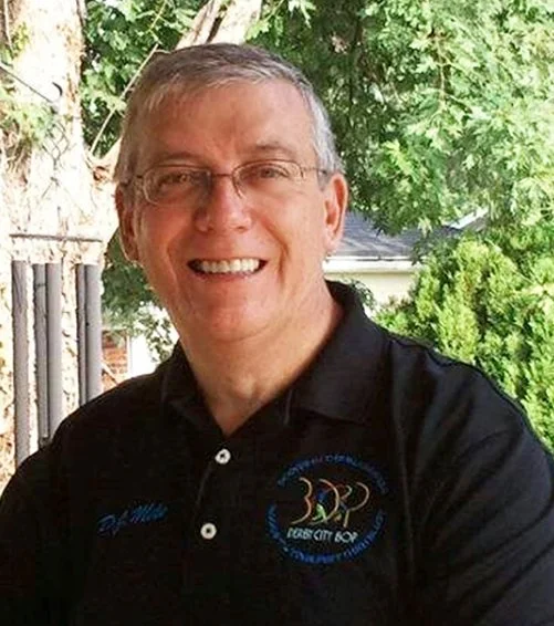 A middle-aged man with glasses smiling outdoors on a sunny day, wearing a black polo shirt with embroidered logos, standing in front of trees and a house.