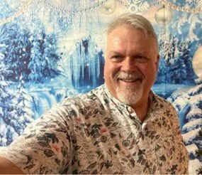 A smiling man with gray hair and beard taking a selfie in front of a snowy winter scene with ice, snow-covered trees, and snowflakes.