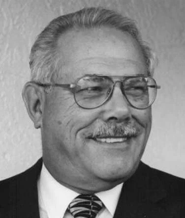A black and white photo of a middle-aged man with glasses, a mustache, and gray hair, smiling.