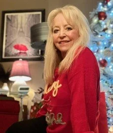 Older woman with long blonde hair smiling, wearing a red sweater, sitting in a decorated living room with a Christmas tree in the background.