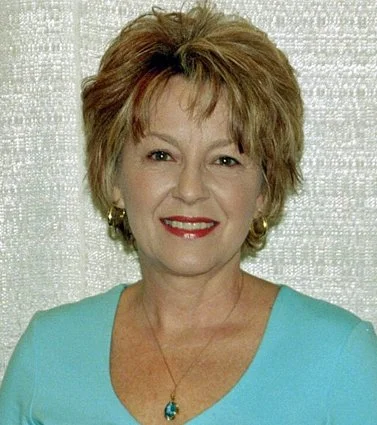 A middle-aged woman with short, wavy, reddish-brown hair smiling and wearing a turquoise top and gold jewelry.