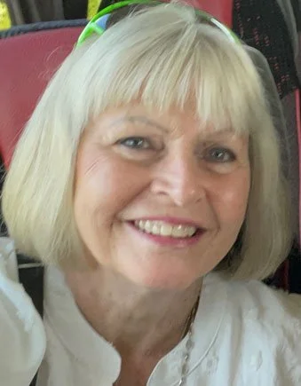 Close-up of a smiling elderly woman with blonde hair and bangs, wearing glasses on her head, in a red vehicle interior.