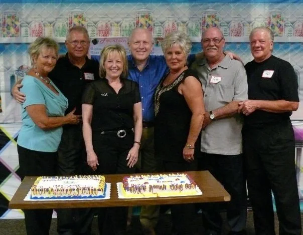 Group of seven adults standing behind a table with two birthday cakes at a celebration. They are smiling and dressed casually, with a birthday banner and decorated backdrop in the background.