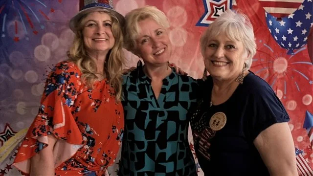 Three women smiling at a celebration with patriotic decorations, including red, white, and blue banners and star-shaped decorations.