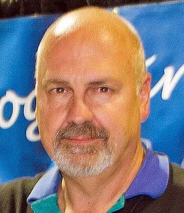 Close-up of a middle-aged man with a bald head and a beard, smiling, standing in front of a blue background with white text.
