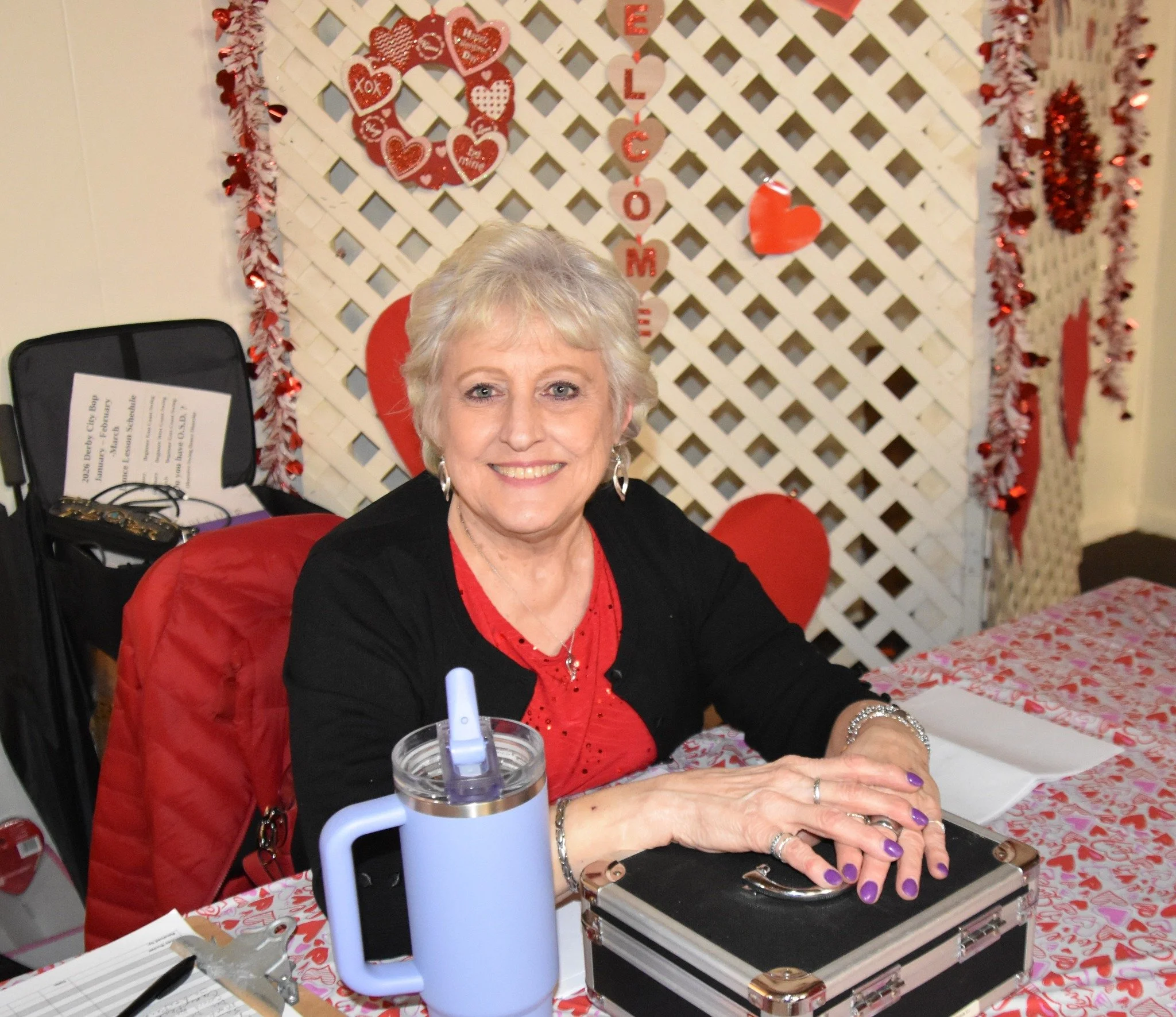 A smiling woman with short gray hair sitting at a table, dressed in a red top and black blazer, with Valentine's Day decorations behind her including hearts and the words 'E LOVE M', and a black case with paperwork on the table, in a festive setting.