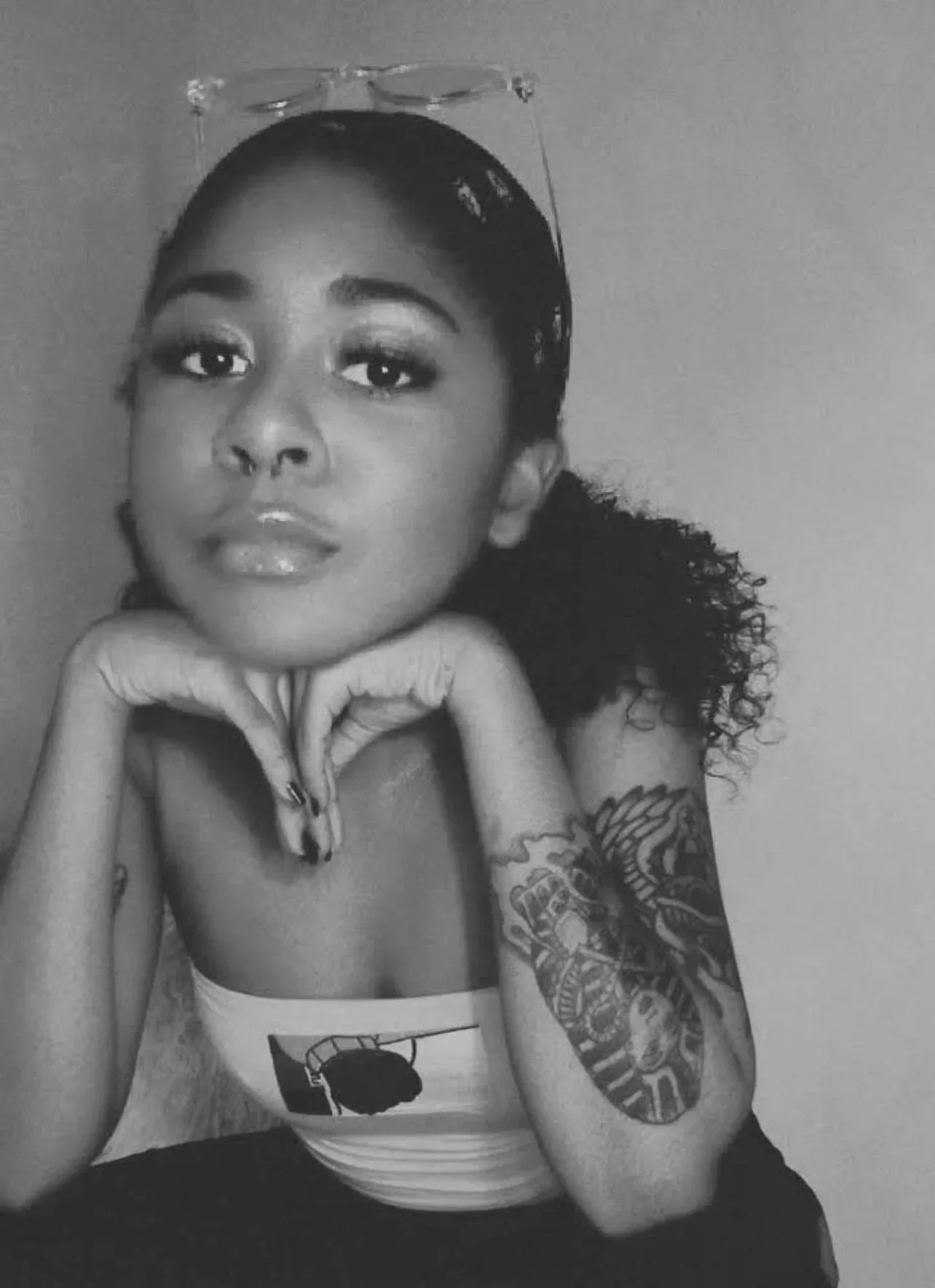Black and white photo of a woman with curly hair, tattoos on her right arm, wearing a tank top, resting her chin on her hands, looking directly at the camera.