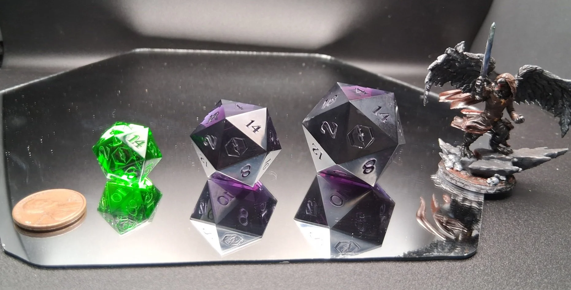 A collection of gaming dice, including a green twenty-sided die, two purple or black twenty-sided dice, and a mini figurine of a winged creature wielding a sword, all on a reflective surface next to a coin.
