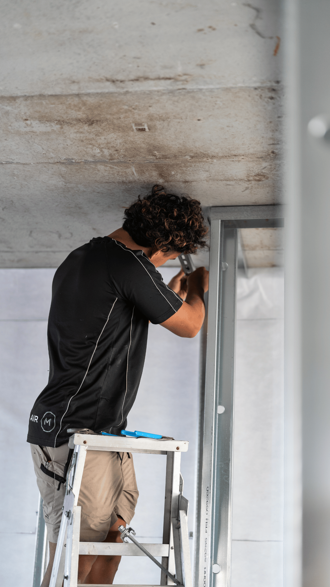 A man in beige shorts and a black T-shirt standing on a ladder and installing or fixing a metal framework against a wall under a concrete ceiling.