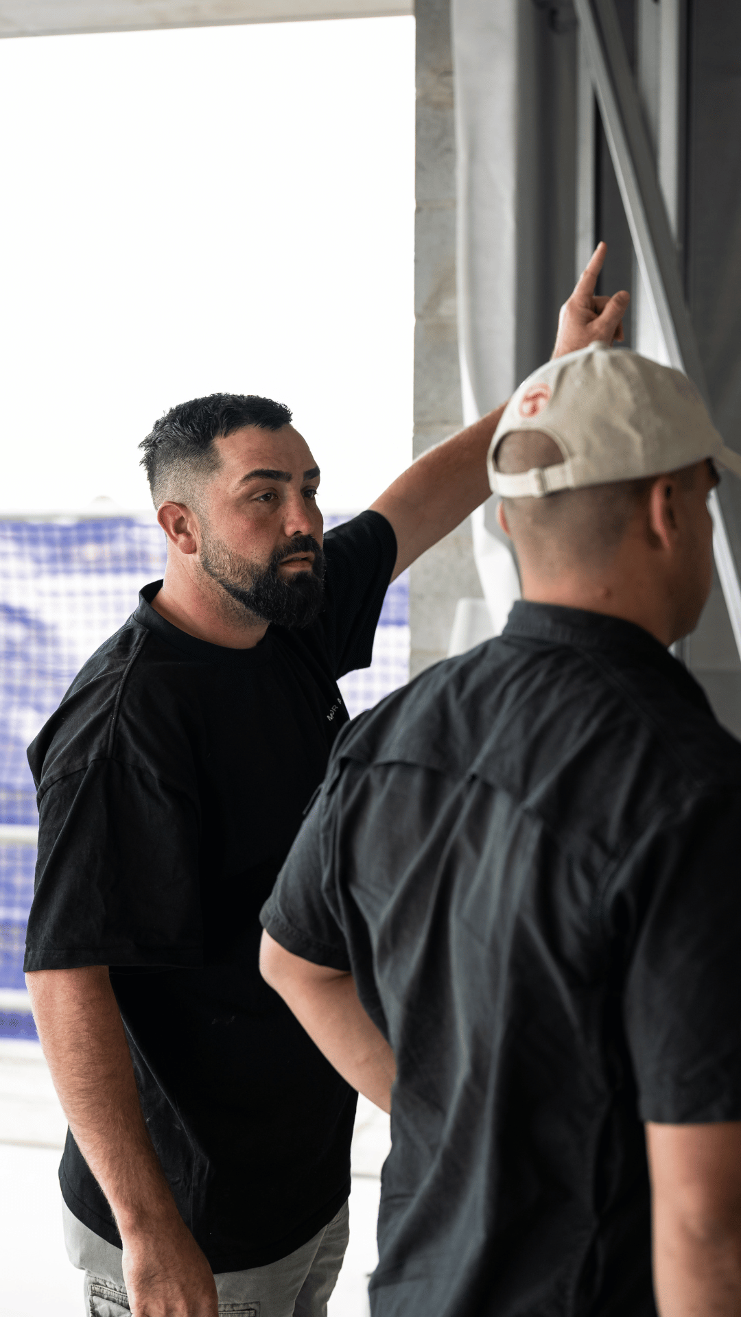 Two men in black shirts, with one pointing at a wall or board in a bright room.