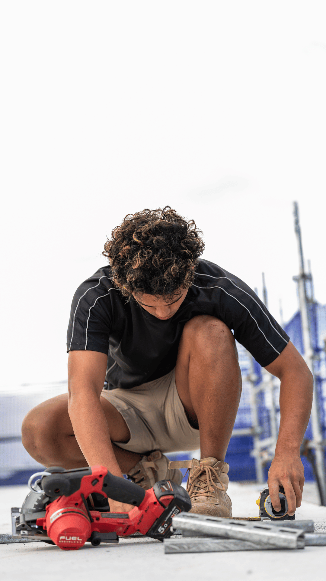 Young man in black shirt and khaki shorts crouching on the ground, working with tools on a construction or renovation project outdoors, with a blue structure and tools in the background.