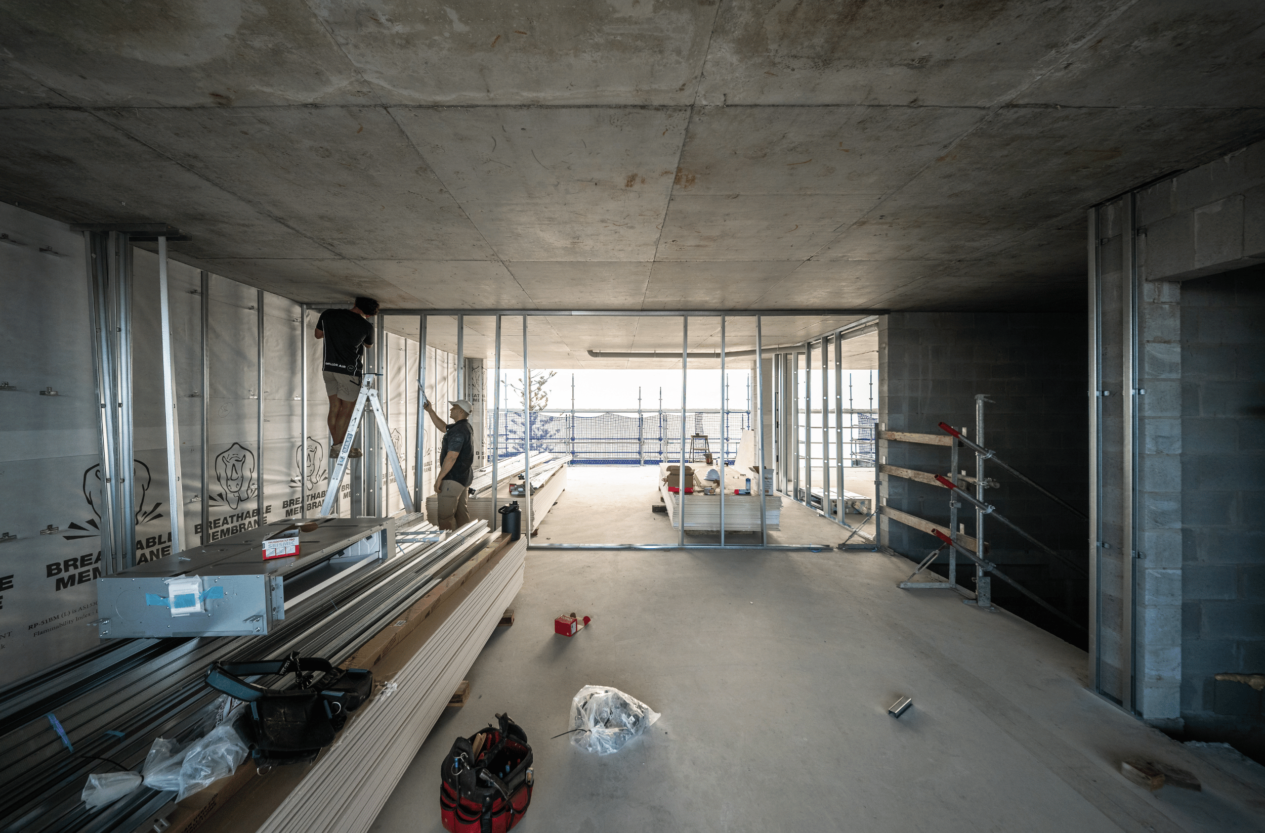 Construction workers installing metal framing in an unfinished building with concrete floors and a large open space.