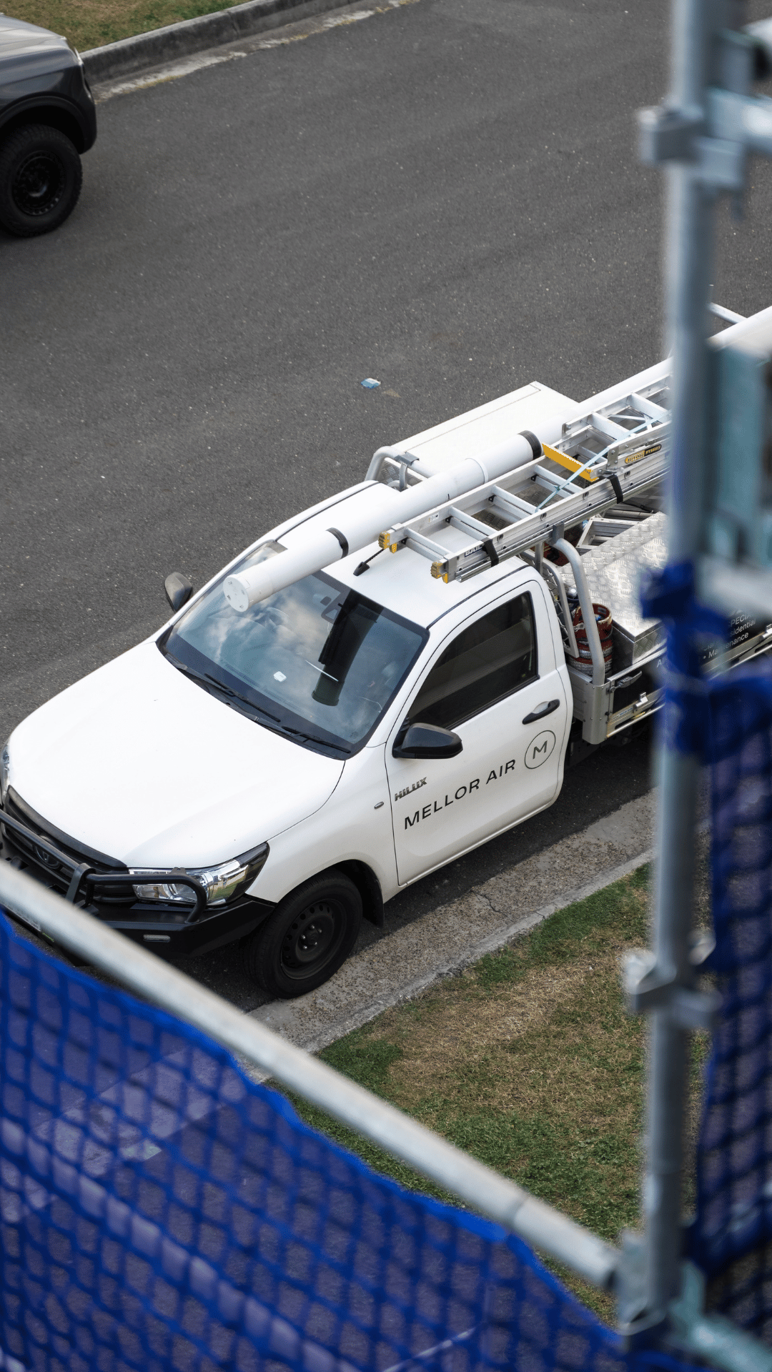 This image shows a white utility truck parked on the side of the road, labeled with 'Mellor Air.' The vehicle is equipped with ladders and other equipment on its bed, and there is a blue safety barrier in the foreground.