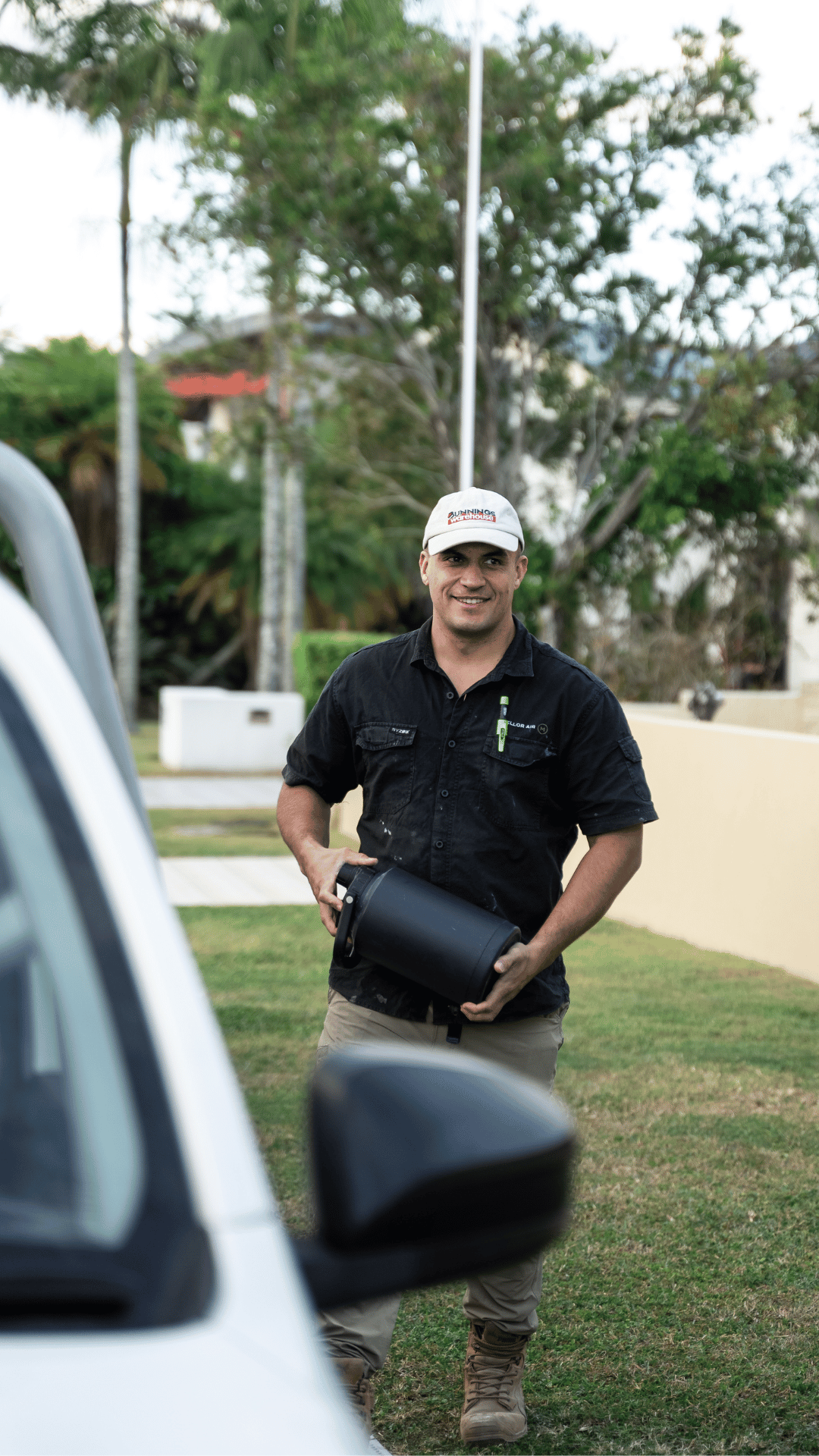 A man standing outdoors holding a black portable speaker, smiling, wearing a white cap with 'Bunnings Warehouse' logo, a black shirt with a green pen in shirt pocket, and tan pants, with trees and a low wall in the background.