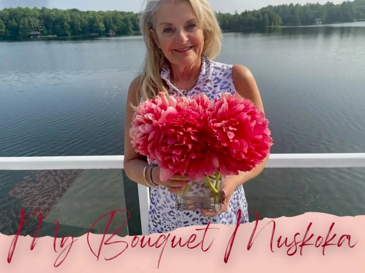 A woman holding a glass vase with pink flowers on a boat or dock, with a lake and trees in the background.