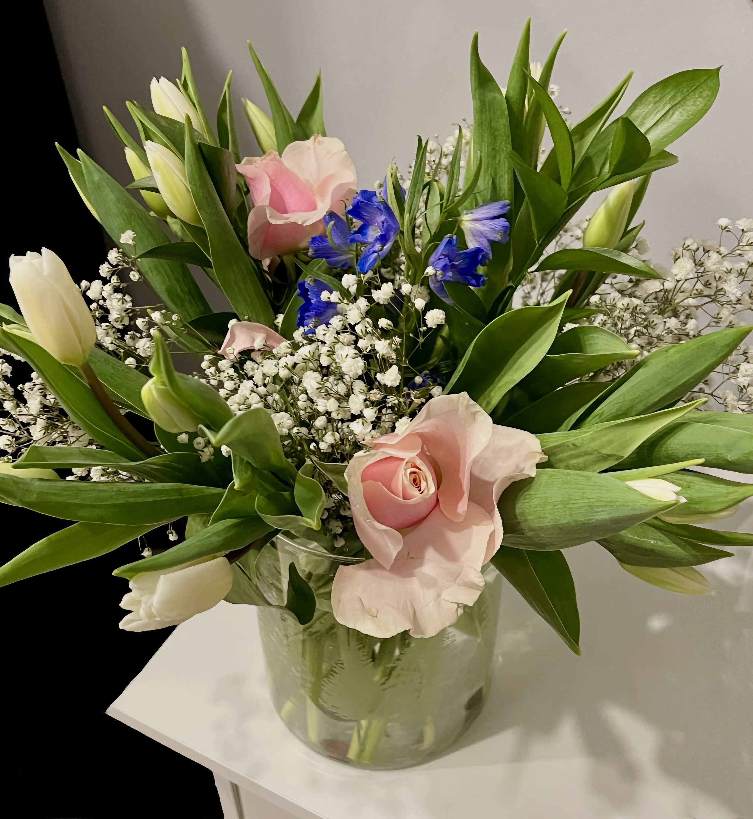A floral arrangement in a translucent green vase on a white surface, featuring pink roses, white lilies, blue irises, small white baby's breath flowers, and abundant green leaves.