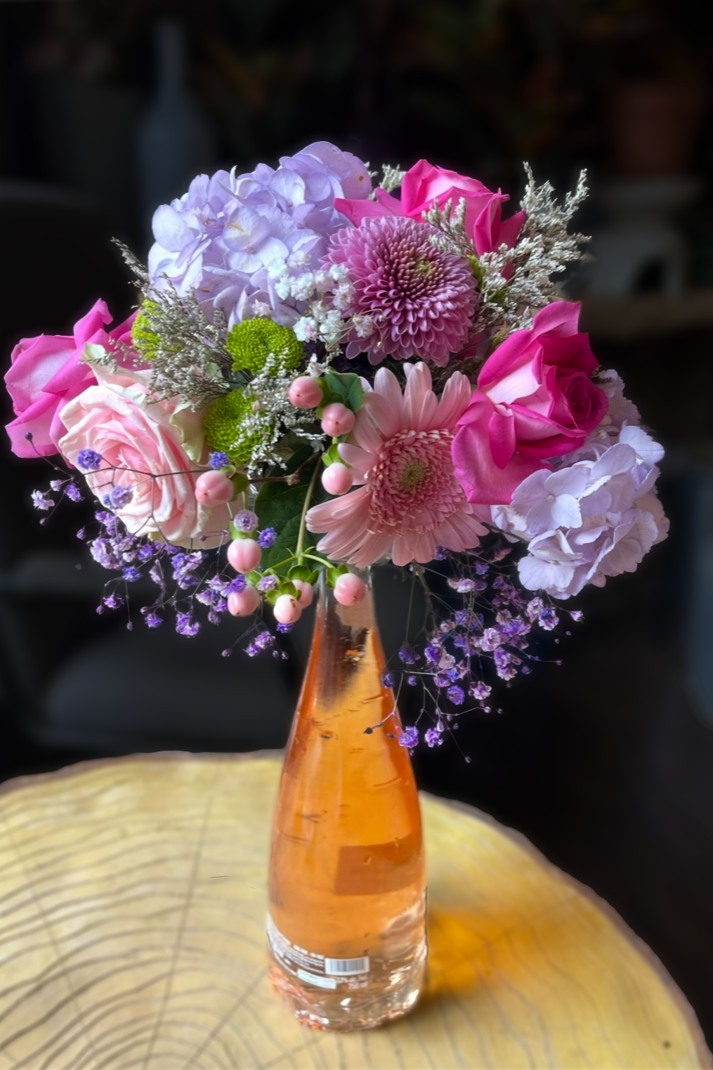 A bouquet of pink, purple, and white flowers in a tall, narrow amber-colored glass vase placed on a wooden surface.