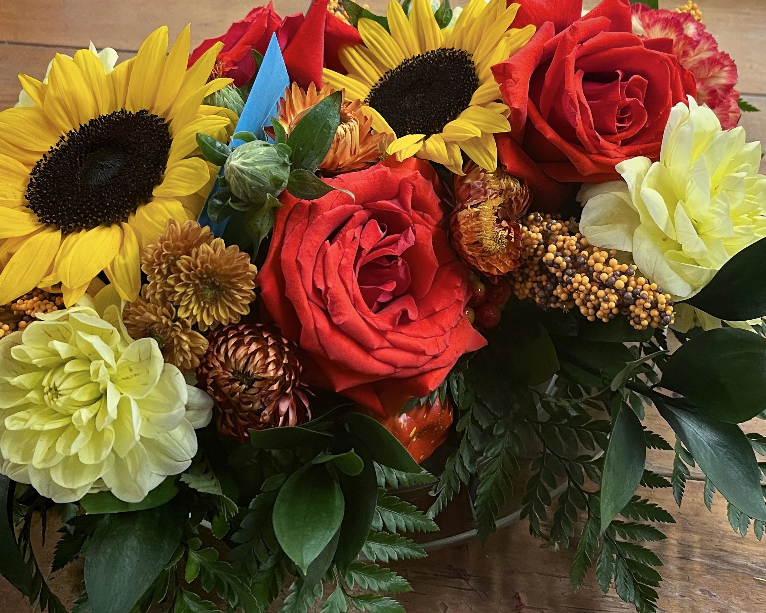 A colorful bouquet of flowers featuring yellow sunflowers, red roses, orange tulips, cream dahlias, and various green leaves, arranged on a wooden surface.