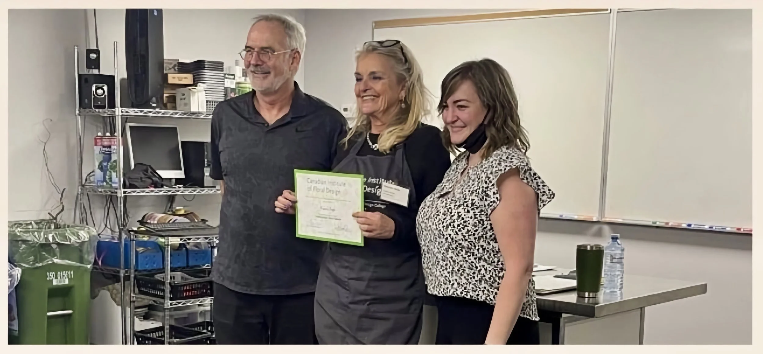 Three people standing together in a room, smiling at the camera. The woman in the middle is holding a certificate. The setting appears to be an office or classroom with shelves, a whiteboard, and various items on tables.