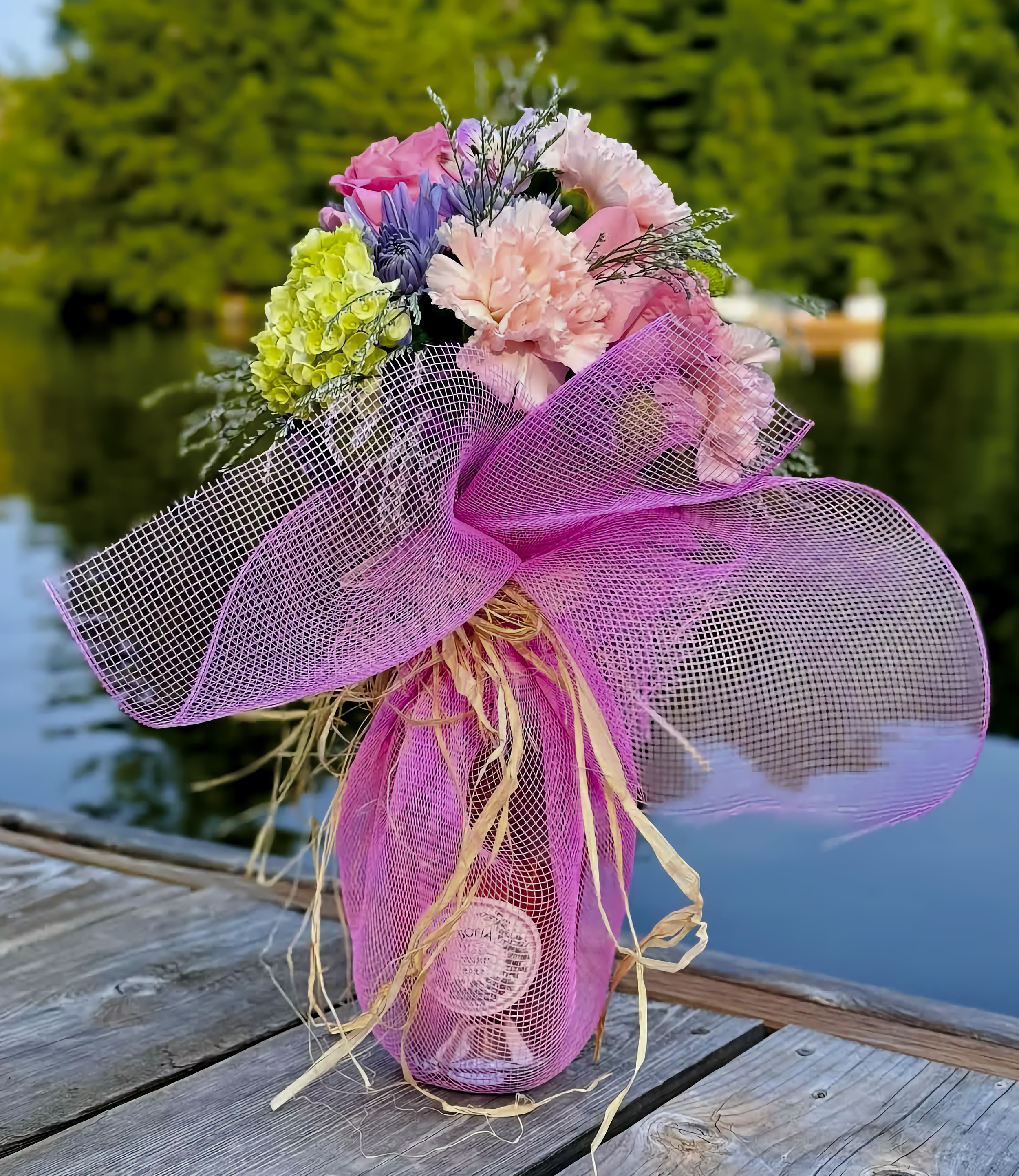 A colorful bouquet of flowers wrapped in pink mesh, placed on a wooden dock by a body of water with a green, tree-lined background.