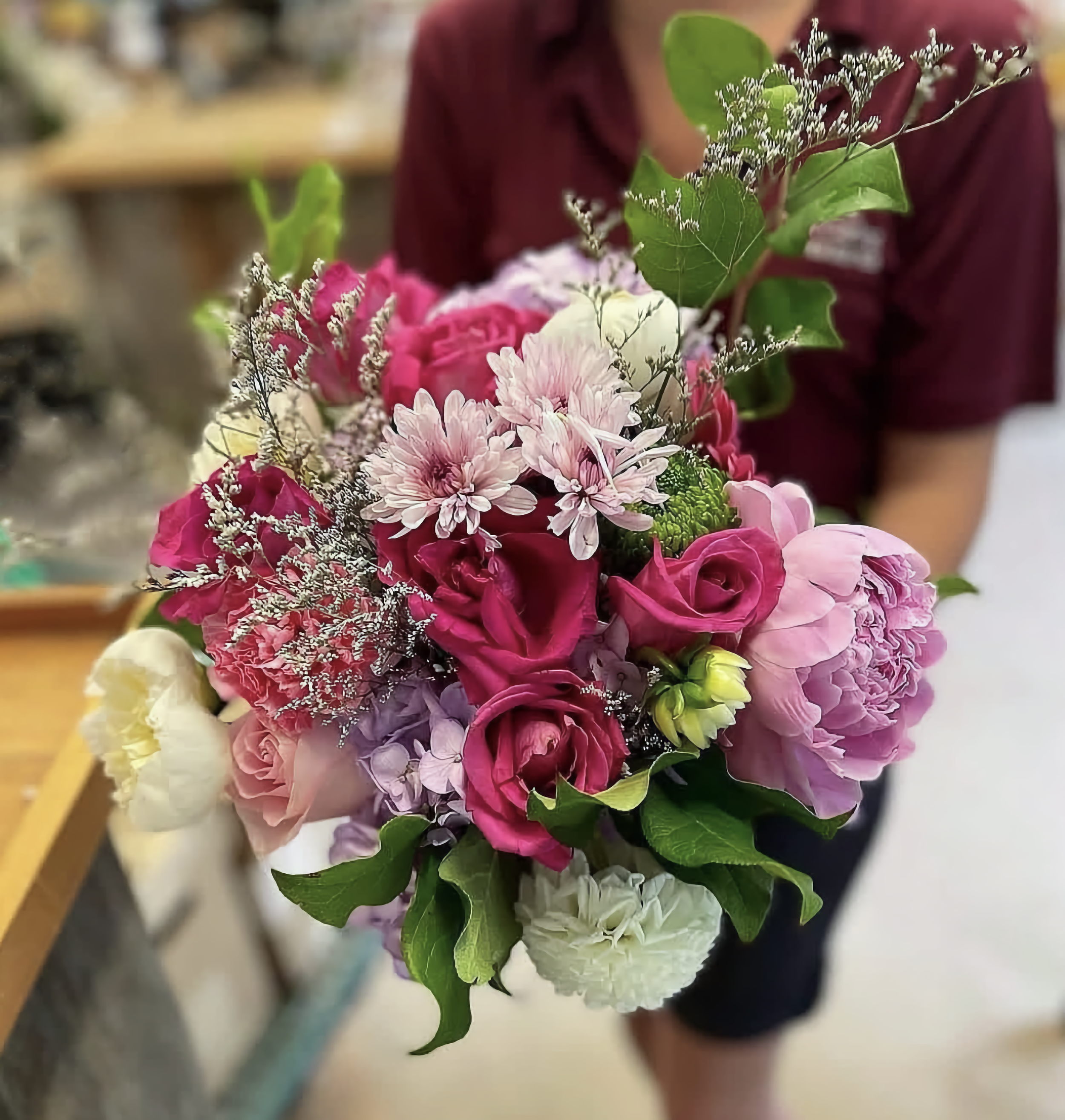 Close-up of a bouquet with pink, white, and purple flowers, and green leaves, held by a person wearing a maroon shirt.