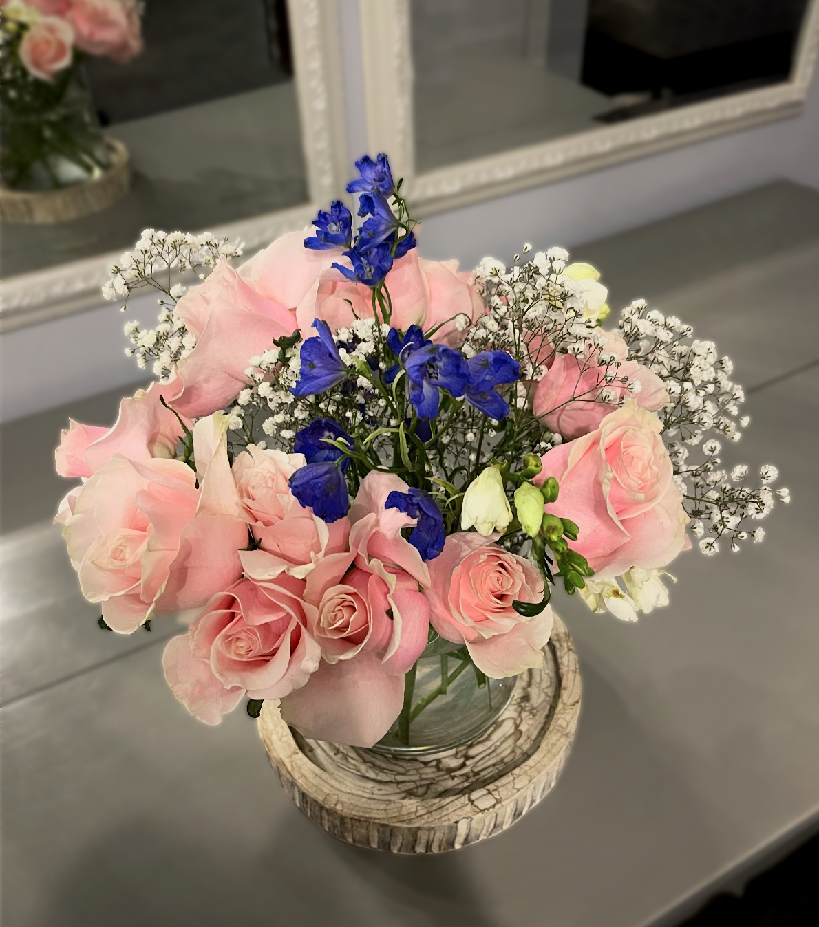 A bouquet of pink roses, blue delphiniums, and white baby's breath in a glass vase on a wooden stand, with a mirror and pink roses in the background.
