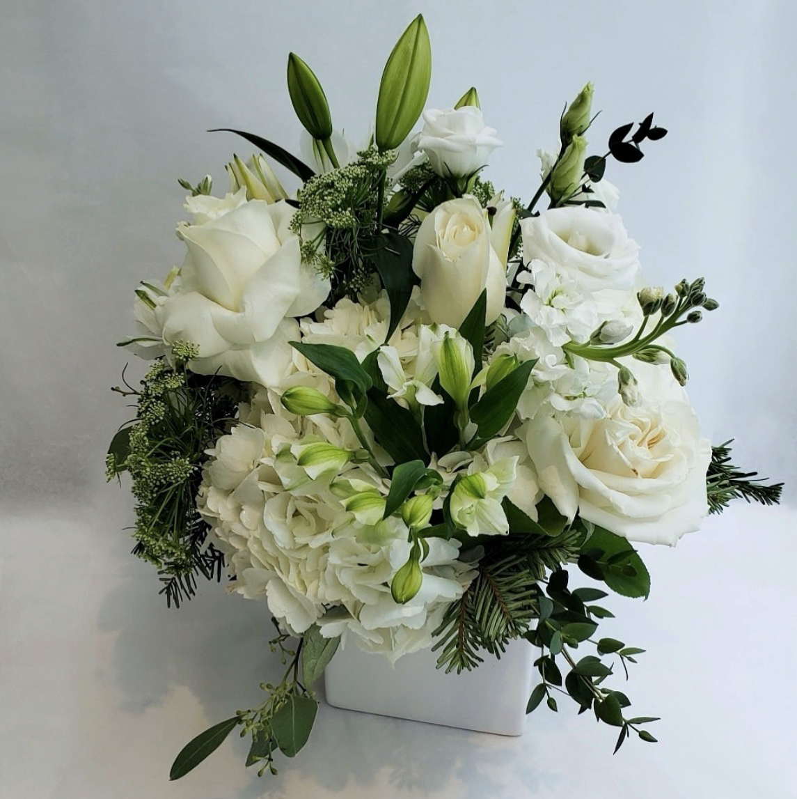 White floral arrangement with roses, lilies, hydrangeas, and greenery in a white square vase.