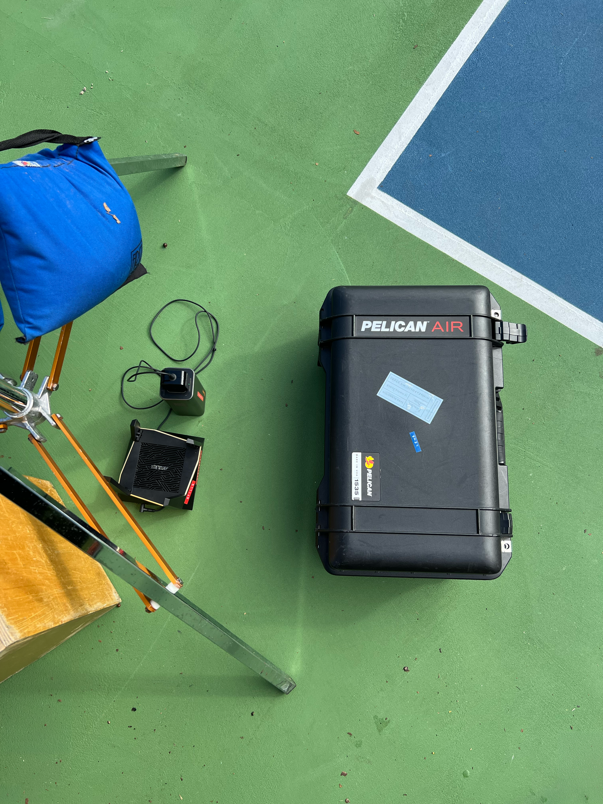 Part of a tennis court with a blue area, green surface, and white boundary lines. Items on the court include a black Pelican Air hard case, a blue bag, a small black electronic device with a cord, a wooden table, and a metal structure.
