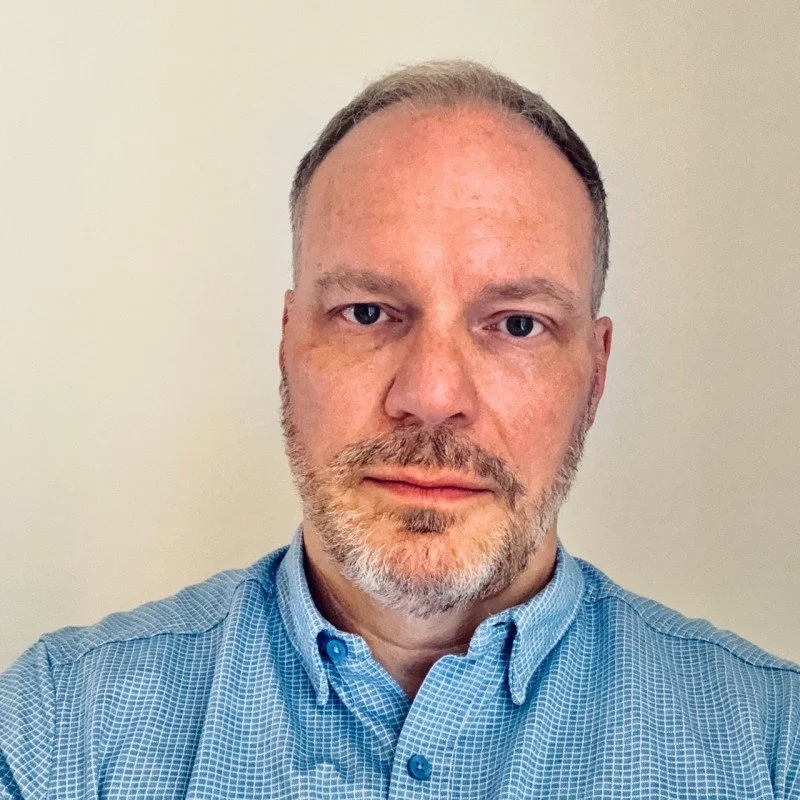 A  man with short hair, a beard, and blue eyes wearing a light blue button-up shirt, standing against a plain light-colored wall.