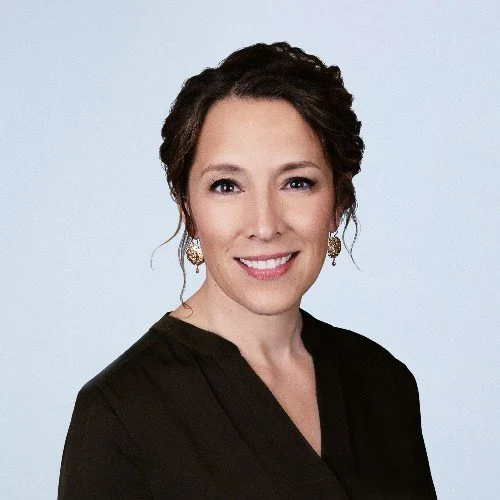 Professional woman with dark curly hair, wearing a black top and earrings, smiling in front of a plain light background.