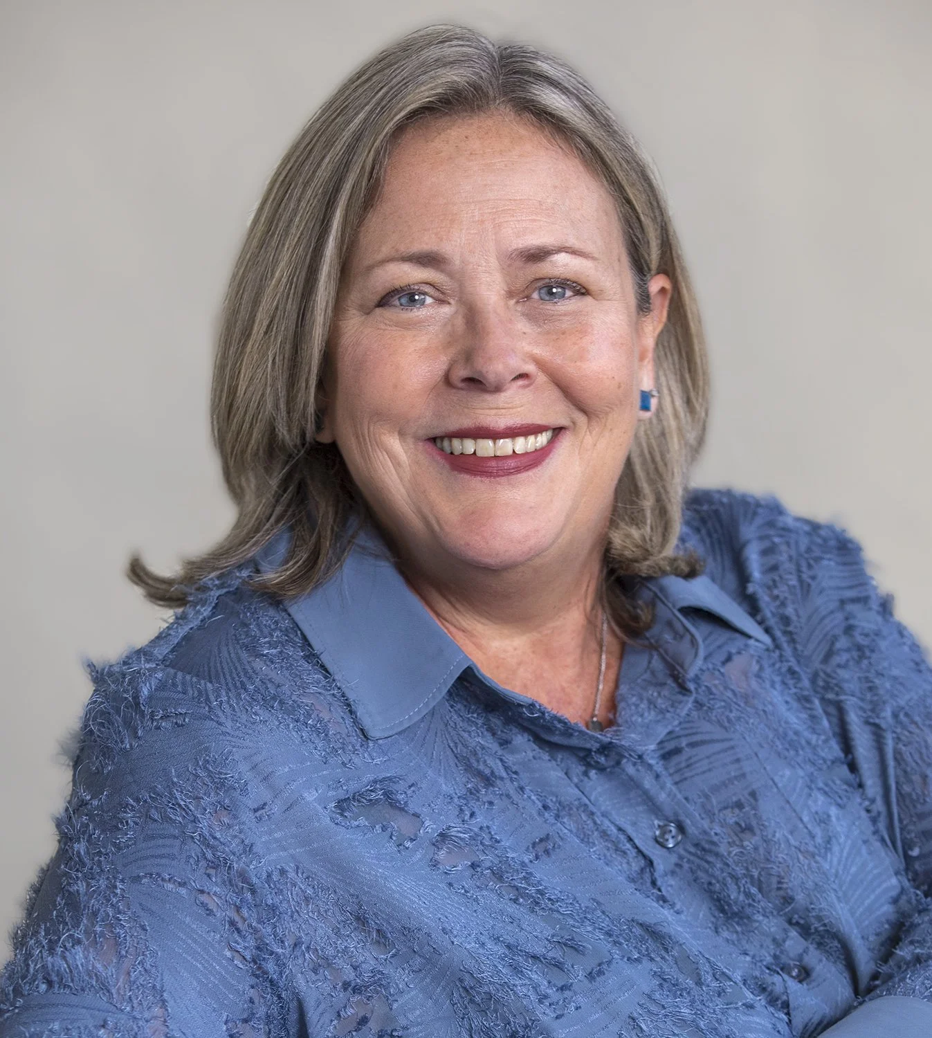 A woman with shoulder-length blond hair, wearing a blue textured blouse, smiling against a neutral background.