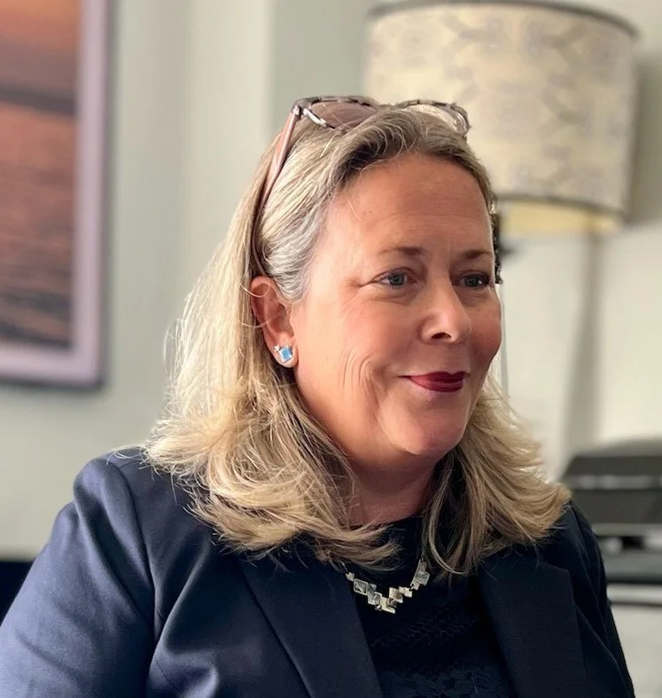A woman with blonde hair, wearing a navy blazer, glasses on her head, and blue earrings, smiling and looking to the side in an indoor office setting.