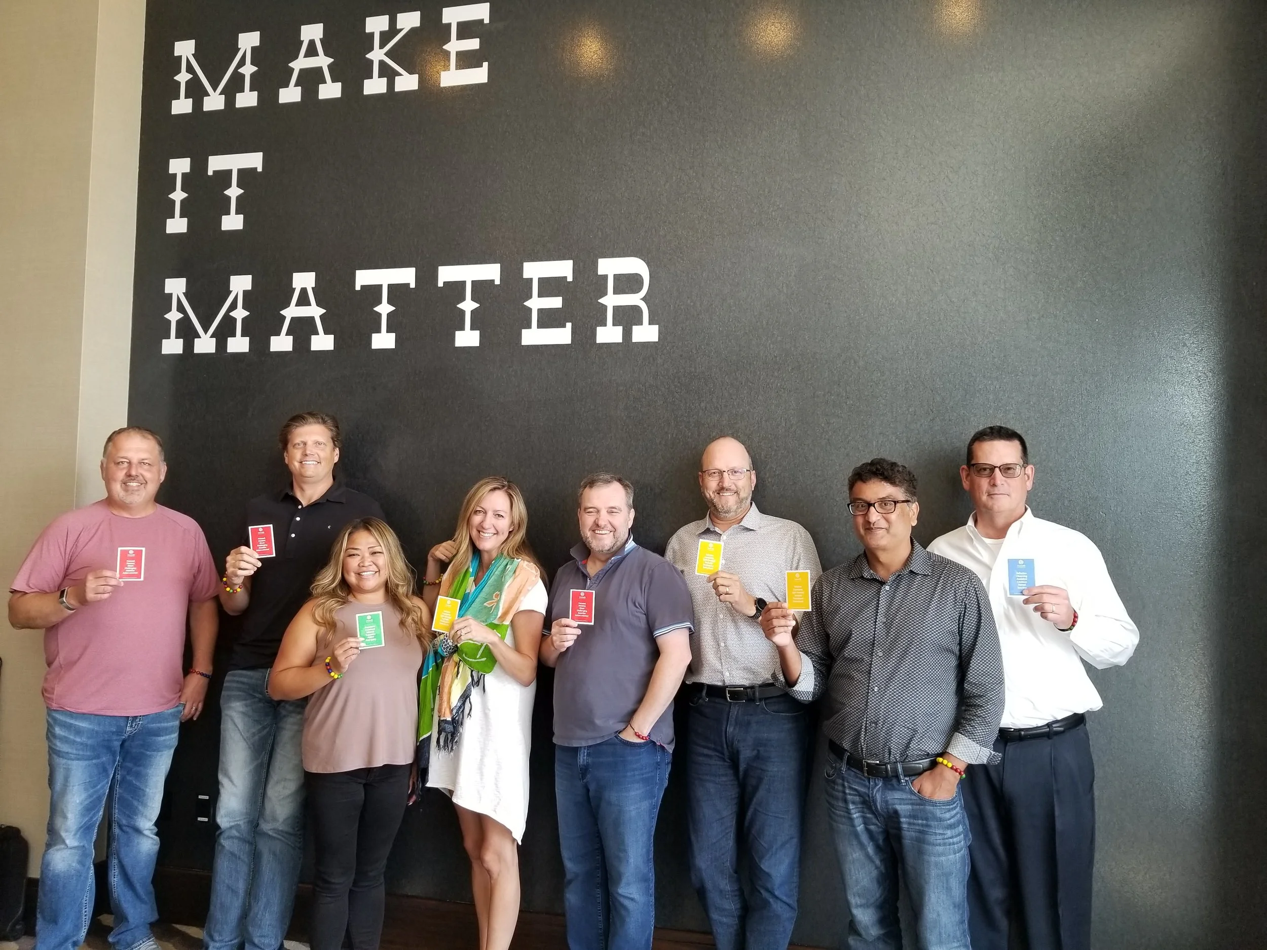 Group of nine people standing together in front of a black wall with white letters that spell 'MAKE IT MATTER'. They are holding colored cards, smiling, and posing for the photo.