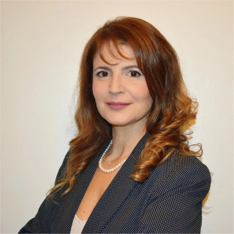A woman with shoulder-length red hair, wearing a black blazer, white top, and a pearl necklace, smiling at the camera against a plain background.