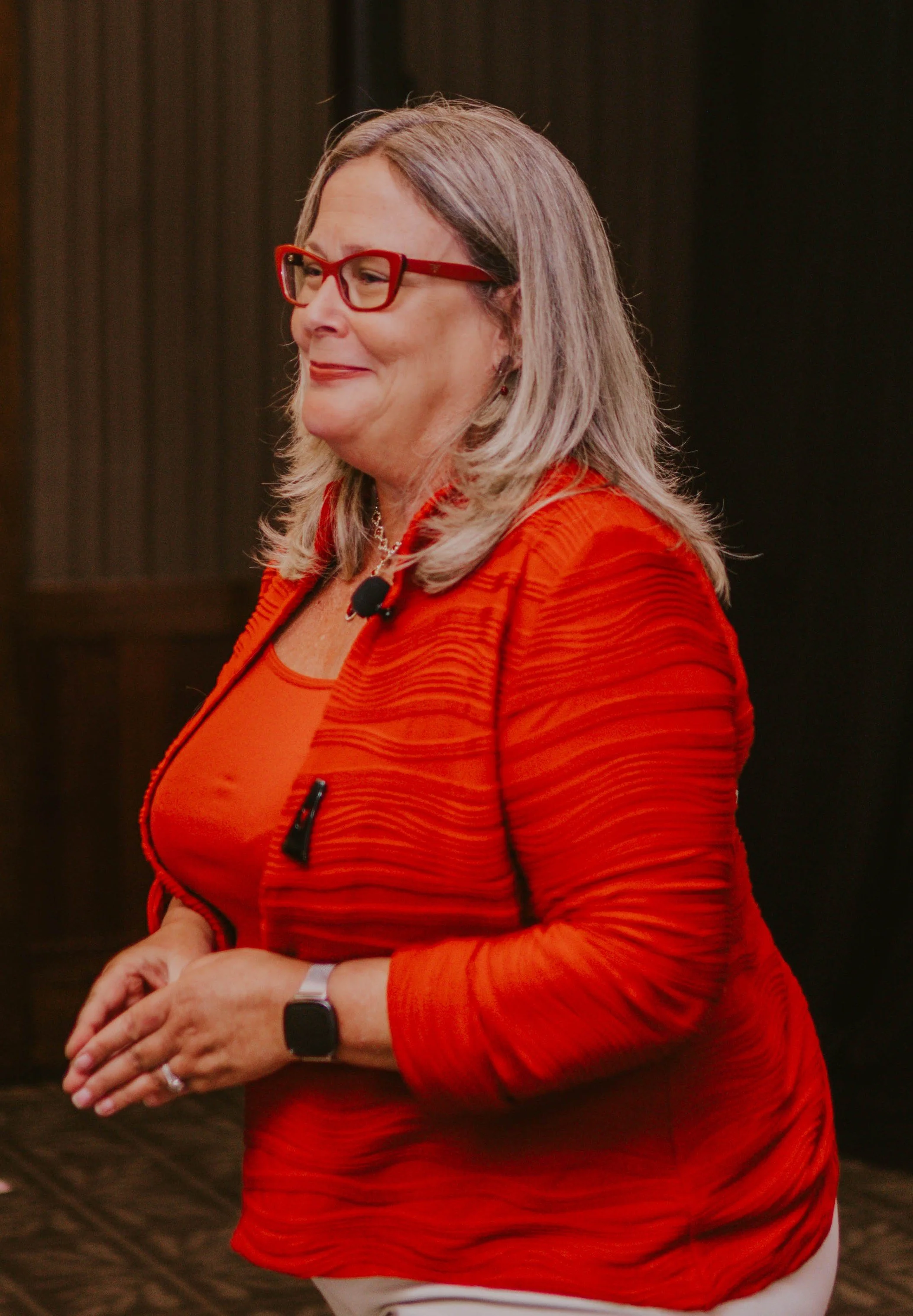 Speaker, Kathy Eastwood, a woman with blond hair and red glasses wearing a red blazer and matching top, standing indoors on a stage addressing an audience.
