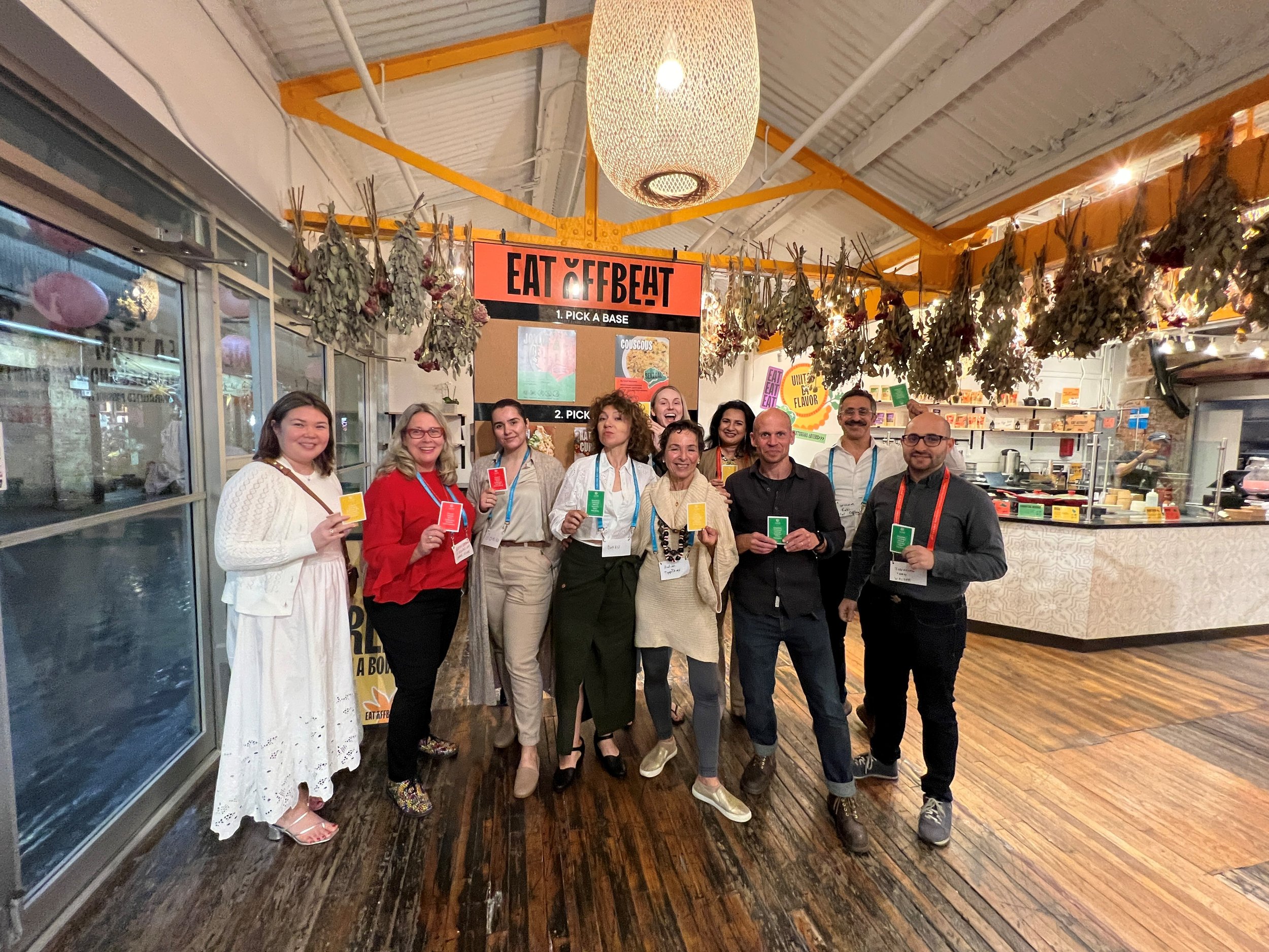 A group of people standing in front of a sign that says 'Eat Offbeat' inside a restaurant or market. The group is smiling and holding colorful cards.