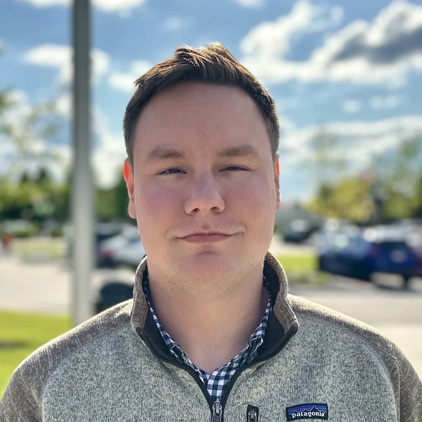 Portrait of a young man outdoors with a blue sky and clouds, wearing a Patagonia fleece jacket and a checkered shirt underneath.